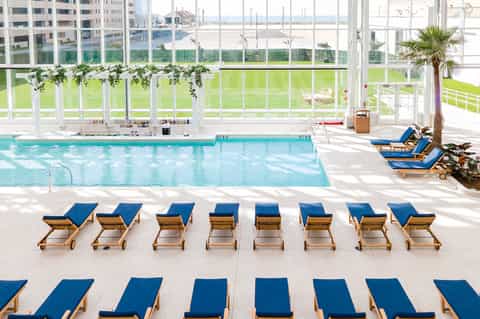 Indoor pool facility with rows of blue lounge chairs, floor-to-ceiling windows, and grassy views
