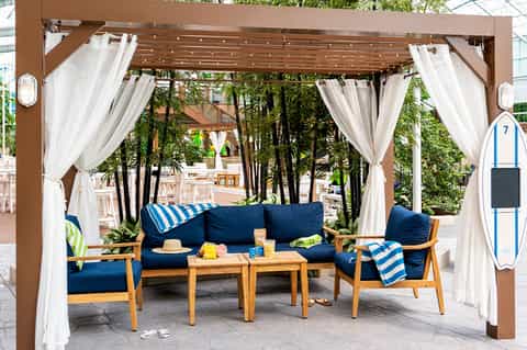 Outdoor cabana with brown pergola, white curtains, blue cushioned seating, and bamboo landscape