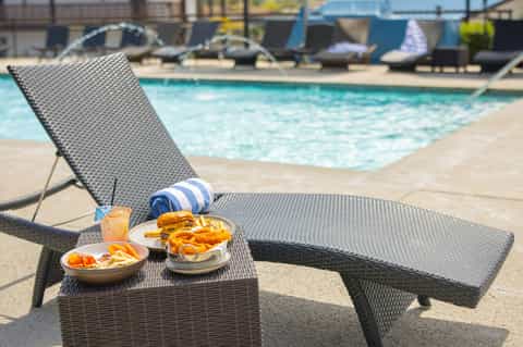 Lounge chair by resort pool with burger, fries, and cocktail on small table, ocean and boats in background