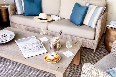 Beige outdoor sofa with blue pillows, wine bottle, and appetizers on a wooden table