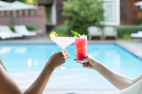 Two hands toasting colorful cocktails poolside with blue swimming pool and resort lounge chairs in background