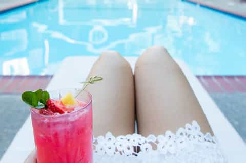 Woman's legs in white shorts holding pink cocktail overlooking resort swimming pool