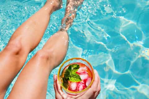Person holding colorful tropical cocktail by pool with legs in turquoise water