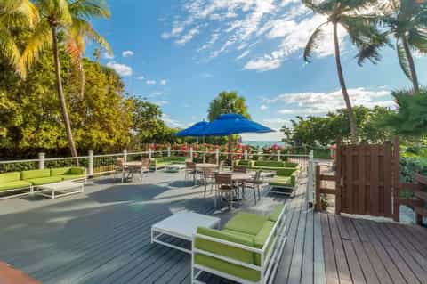 Oceanfront deck with green lounge chairs, blue umbrella, palm trees, and water views