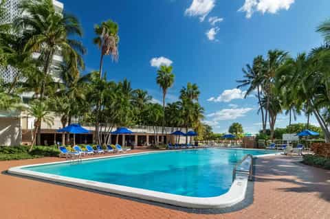 Resort pool surrounded by palm trees, blue umbrellas, lounge chairs, and tropical landscaping