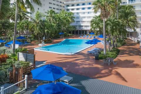 Large resort pool surrounded by palm trees, blue umbrellas, and high-rise hotel building