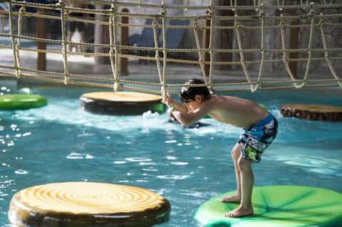 Young boy playing on inflatable platforms in indoor swimming pool with rope obstacle course