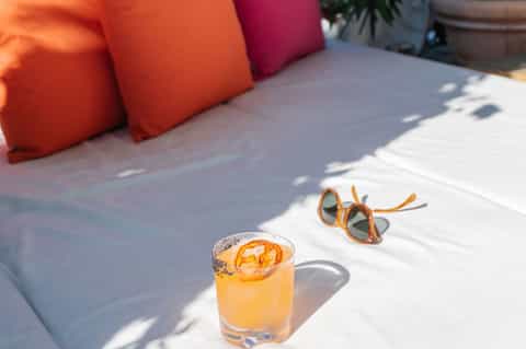Poolside lounger with colorful pillows, orange cocktail, and sunglasses on white cushion