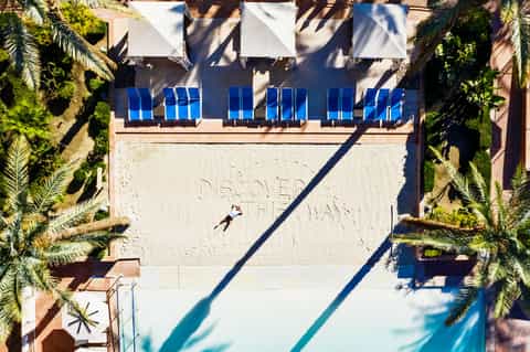 Aerial view of resort pool with blue loungers, white pergolas, and palm trees, guest relaxing on sand