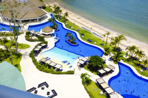 Aerial view of resort with curved blue pools, beach access, palm trees, and lounge chairs