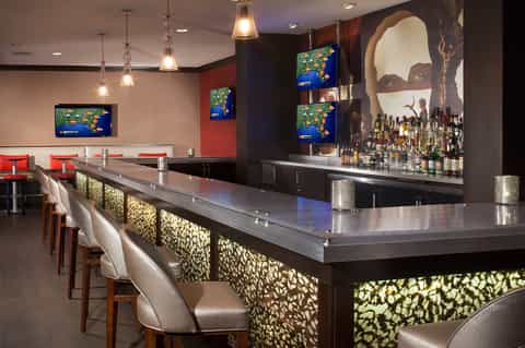 Modern hotel bar with dark wood finishes, illuminated back bar, flat-screen TVs, and geometric patterned panels