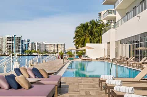 Resort waterfront pool deck with pink loungers, infinity pool, palm trees, and waterfront cityscape views