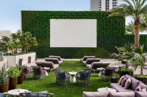 Modern rooftop lounge with artificial grass, lounge seating, purple accents, palm trees, and outdoor screen