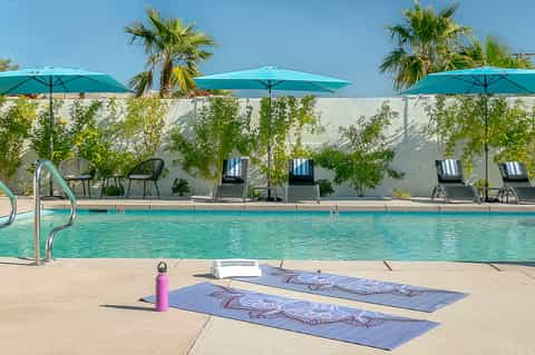 Outdoor resort pool with turquoise umbrellas, palm trees, white wall backdrop, and yoga mats