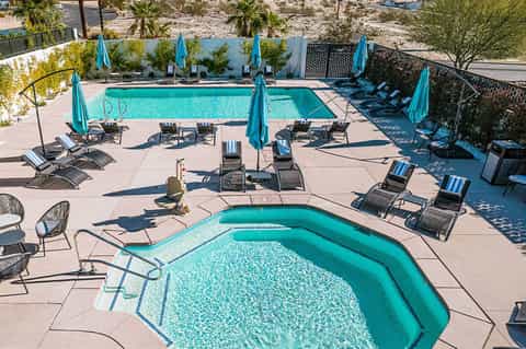 Resort pool area with turquoise water, lounge chairs, blue umbrellas, and desert landscape