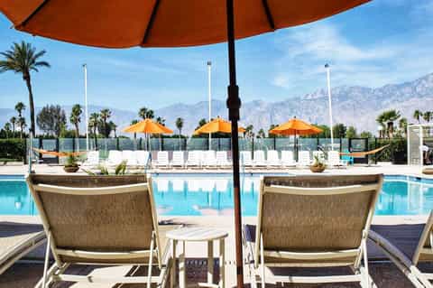 Luxury resort pool with orange umbrellas, mountain views, palm trees, and lounging chairs under shade structures
