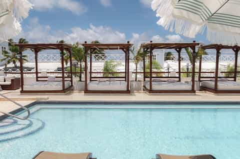 Pool area with wooden cabanas, striped umbrellas, loungers, and beachfront building