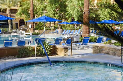 Resort pool area with blue umbrellas, lounge chairs, and tropical palm trees surrounding