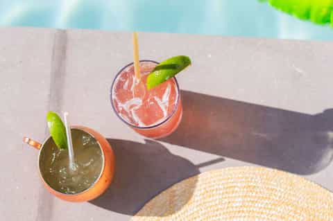 Two colorful cocktails on pool deck ledge with straw hat, clear blue water visible in background