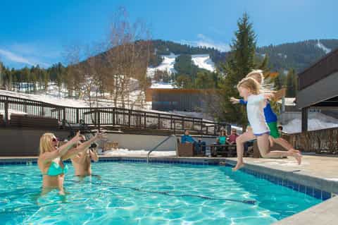 Child jumping into turquoise pool with snowy mountains visible in background and spectators poolside