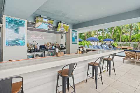 Beach bar counter with bottles and view of pool, umbrellas, and palm trees