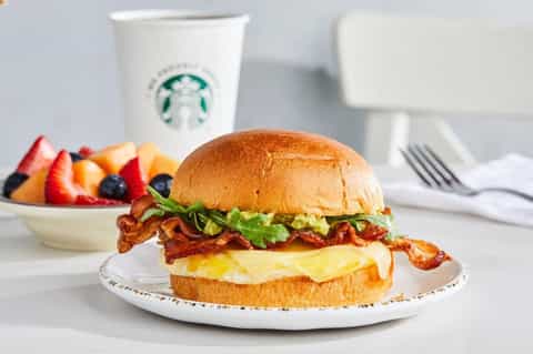 Breakfast sandwich with bacon and egg beside fruit bowl and coffee cup.