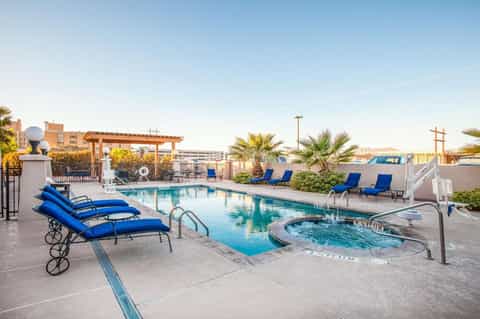Rooftop pool with blue lounge chairs, hot tub, palm trees, and city buildings in background