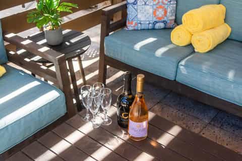 Sunny outdoor patio with blue lounge cushions, wine bottles, glasses, and rolled yellow towels