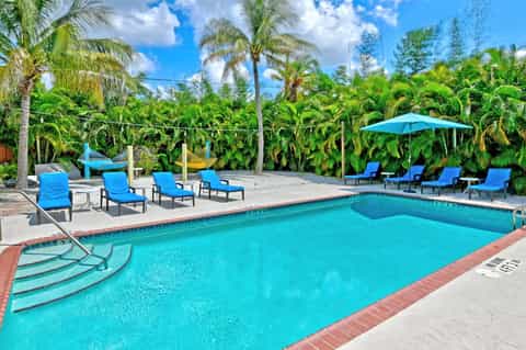 Bright blue pool with loungers, palm trees, tropical landscaping, and blue umbrellas