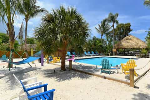 Colorful resort pool area with bright lounge chairs, hammocks, thatched cabana, palm trees, and sandy beach
