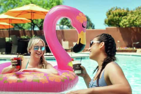Two women on pink flamingo pool float with tropical drinks at sunny resort pool