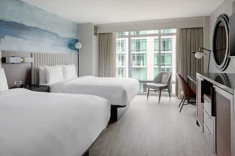 Contemporary hotel bedroom with two queen beds, blue accent wall art, floor-to-ceiling windows, and modern furnishings