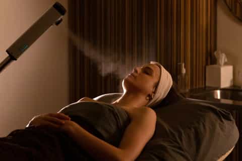 Woman receiving spa steam treatment in dimly lit wellness room with gold curtains