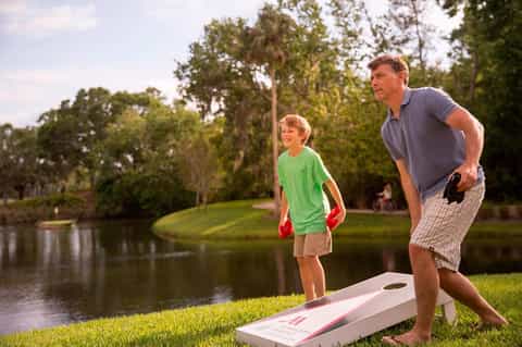 Father and son playing cornhole by a pond surrounded by green lawn and trees