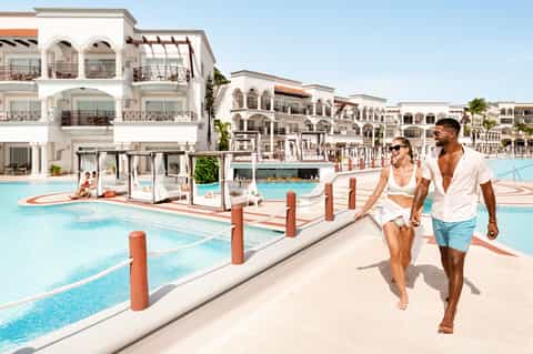 Couple strolling along resort pool deck with white Mediterranean-style buildings and cabanas