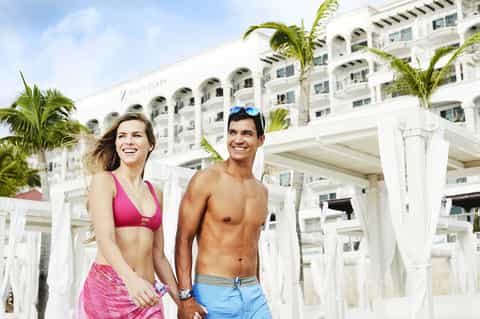 Couple in beachwear standing at resort cabanas with white building and palm trees behind