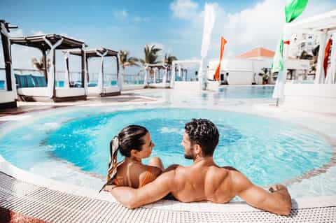 Resort pool with couple in water, cabanas, colorful flags, white lounge beds, and palm tree landscaping