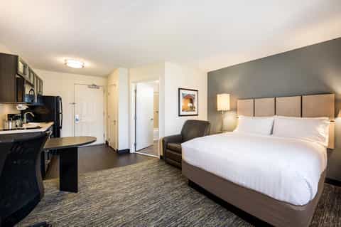 Modern hotel suite with queen bed, kitchenette, dark wood accents, and contemporary furnishings