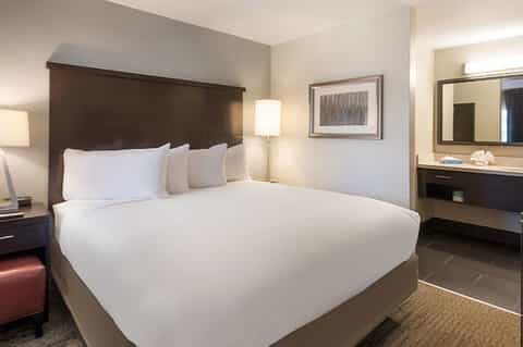 Hotel bedroom with dark wood headboard, white bedding, vanity mirror, and modern furnishings