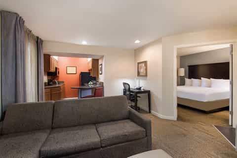 Spacious hotel suite with sofa, kitchenette with orange accent wall, and bedroom visible through doorway