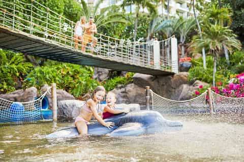 Children playing in shallow resort water feature with suspension bridge and tropical plants overhead