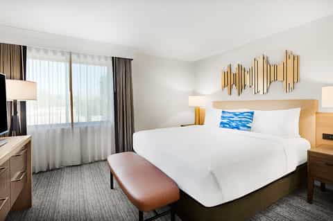 Hotel bedroom with queen bed, white bedding, ocean view balcony, and modern wood furnishings