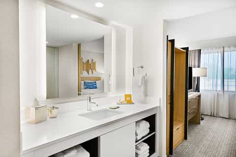 Modern hotel bathroom with white vanity, illuminated mirror, ocean view, and bedroom beyond