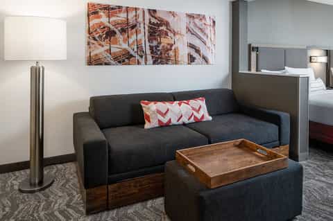 Modern hotel suite with dark sofa, wooden ottoman, and view of bedroom beyond