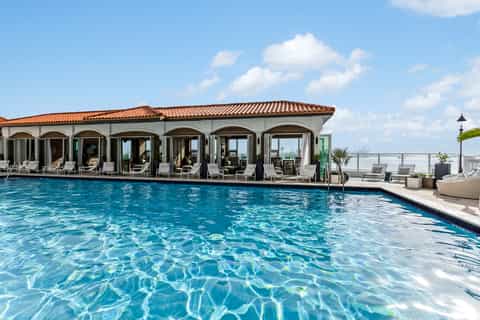 Resort pool with arched cabana building, lounge chairs, and ocean backdrop on horizon