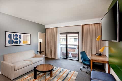 Modern hotel suite with beige sofa, desk, blue accent chair, and balcony
