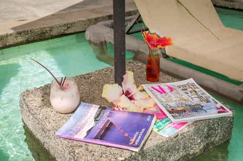 Poolside drinks and magazines on stone table with tropical flowers and turquoise water
