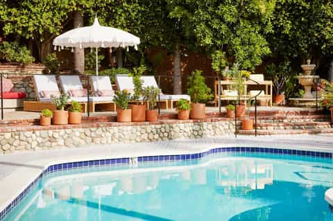 Luxury resort pool with white umbrella, lounge chairs with pink cushions, landscaped stone wall, and mature greenery backdrop