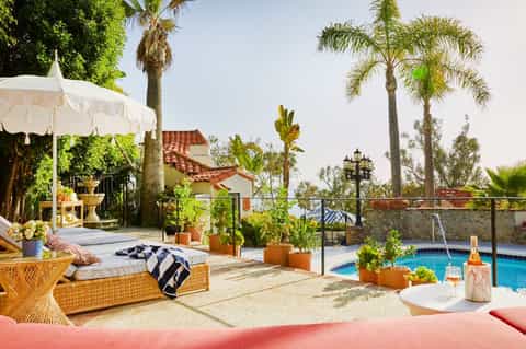 Mediterranean-style pool area with lounge seating, white umbrella, palm trees, and Spanish architecture
