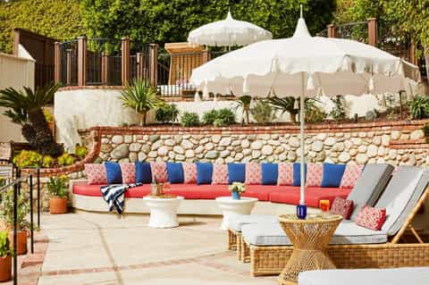 Luxury outdoor lounge with red cushioned seating, coral patterned pillows, white umbrellas, wicker side tables, and stone walls